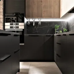 Nowoczesna kuchnia w ciemnej tonacji z blatem imitującym marmur, oświetleniem LED podszafkowym i elementami drewnianymi, widok na ścianę z cegły i wiszące kieliszki.