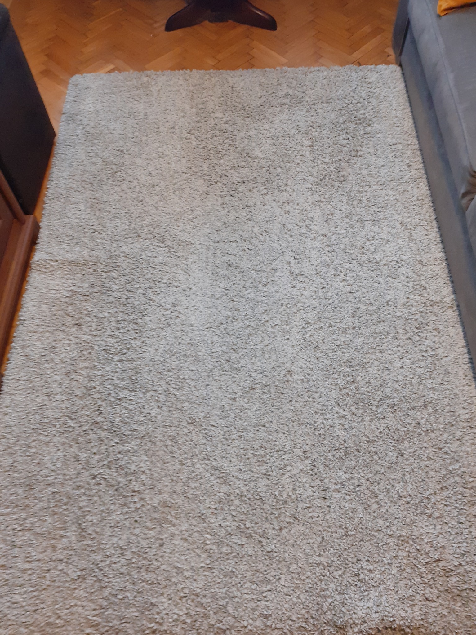 Beżowy dywan shaggy leżący na parkiecie, częściowo przykryty przez szarą kanapę i ciemny mebel.