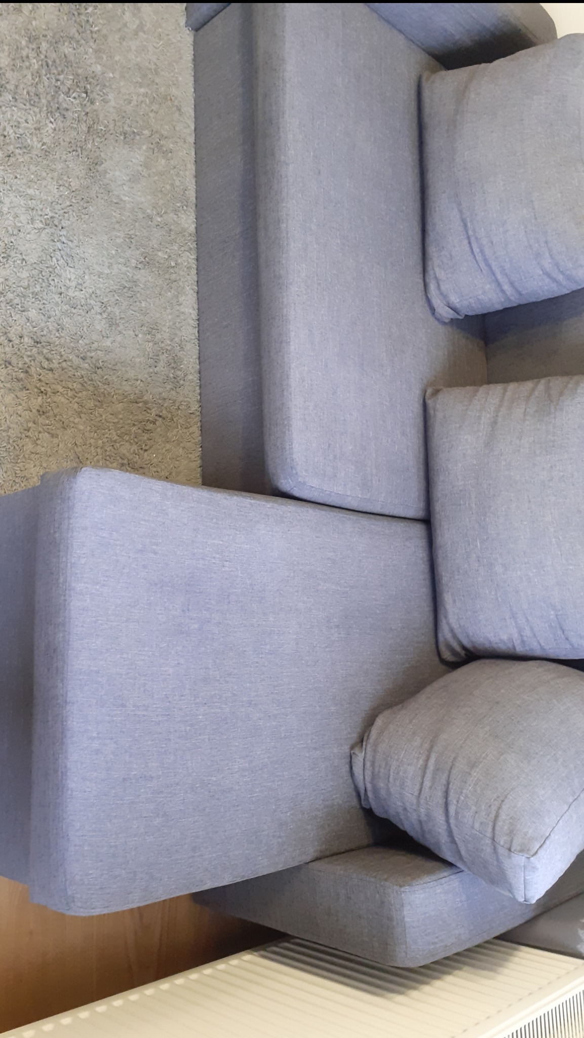 Fragment narożnika tapicerowanego jasnoniebieską tkaniną, widok z góry, obok dywan z krótkim włosiem i grzejnik.
