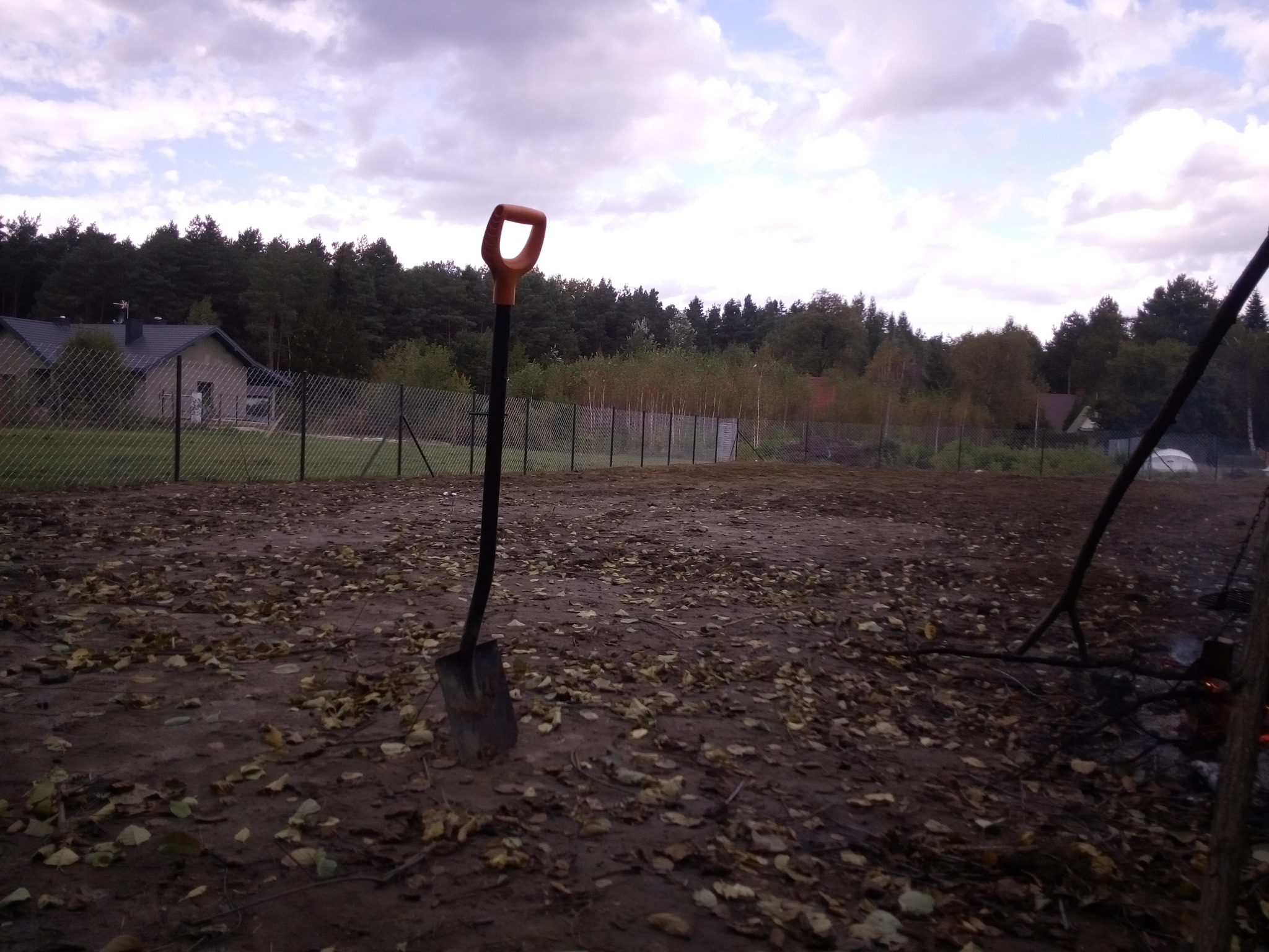 Przygotowany teren pod budowę: wykarczowane drzewa, wyrównana ziemia, wbita łopata w ziemię, w tle siatka metalowa i zabudowania.