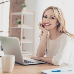 Uśmiechnięta blondynka opiera podbródek na dłoni, siedząc przy biurku z laptopem, kubkiem i przyborami do pisania w jasnym biurze.
