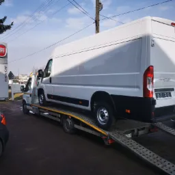 Biały furgon Fiat Ducato na platformie niskopodwoziowej przy salonie samochodowym Fiat Elwa-Car, widoczne najazdy platformy i częściowo zaparkowany szary samochód osobowy.