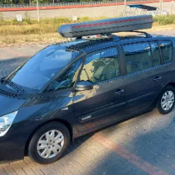 Ciemnoszary van Renault Espace z boxem dachowym na bagażniku, zaparkowany na brukowanej ulicy w słoneczny dzień.