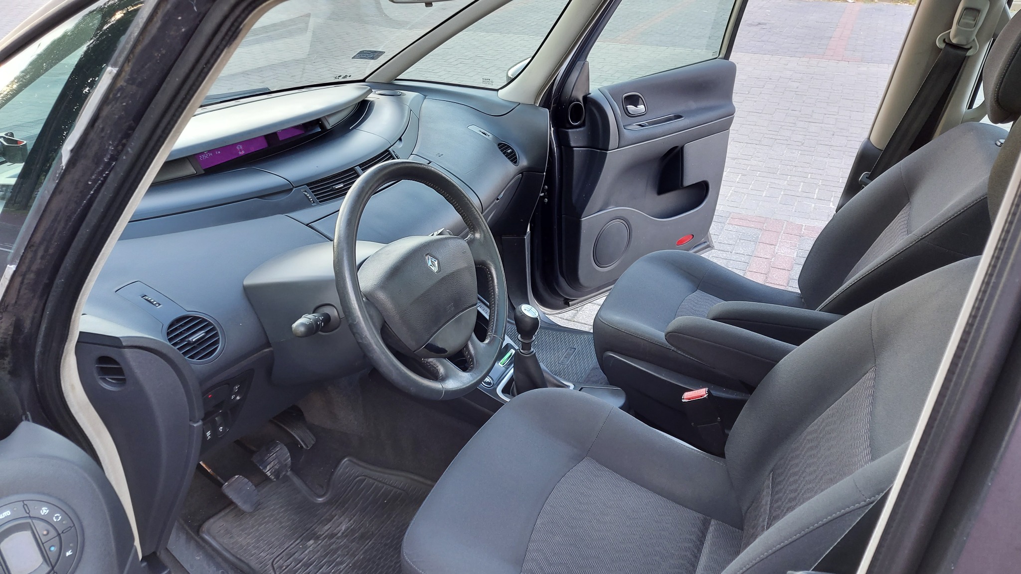 Wnętrze minivana z widocznymi fotelami, deską rozdzielczą i otwartymi drzwiami pasażera, z logo Renault na kierownicy.