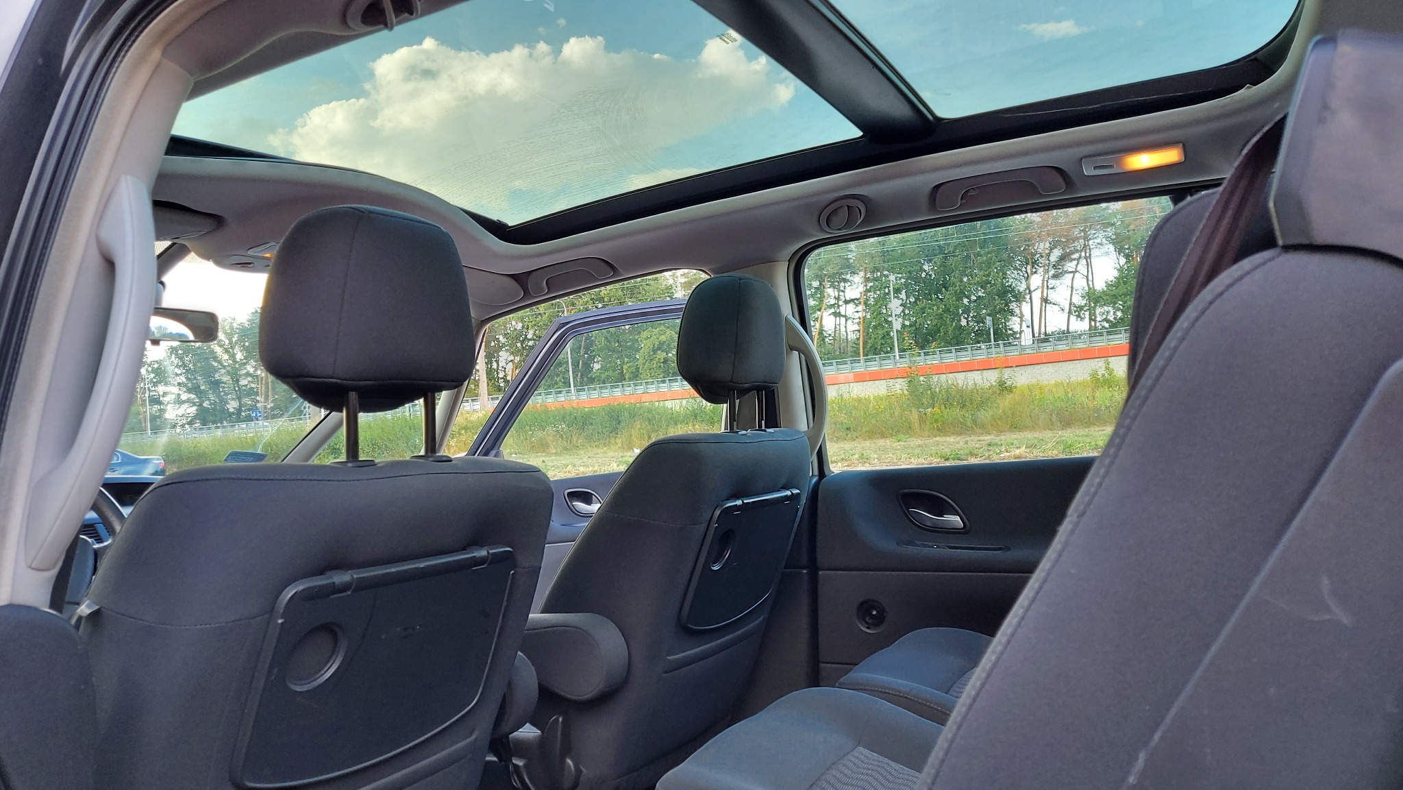 Wnętrze samochodu z panoramicznym dachem, widok z tylnych siedzeń na przednie fotele z rozkładanymi stolikami, otwarte drzwi boczne i widok na zieleń za oknem.