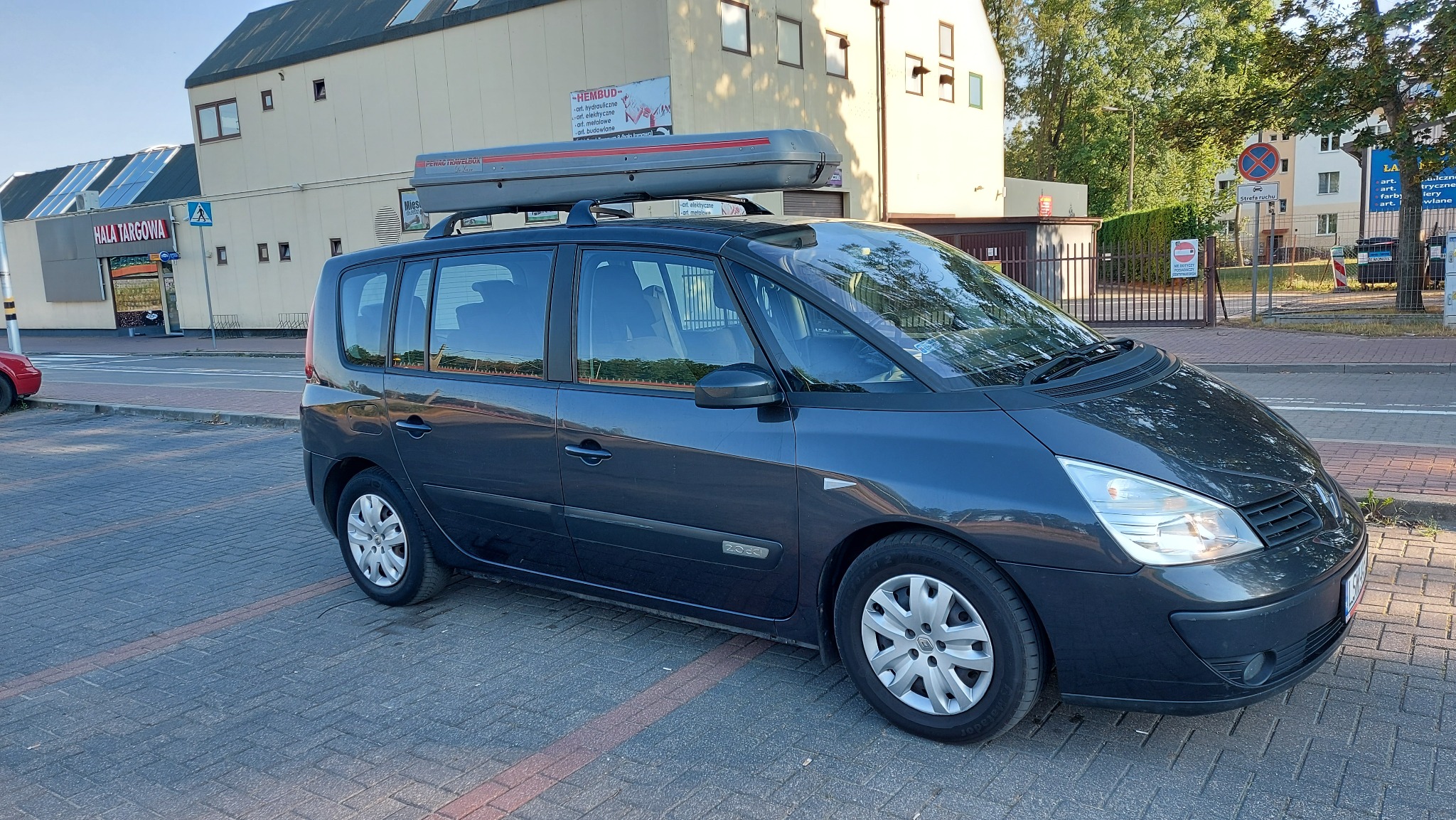Ciemny minivan z bagażnikiem dachowym na parkingu w Świdniku, widok z boku, w tle budynek hali targowej i budynki mieszkalne.