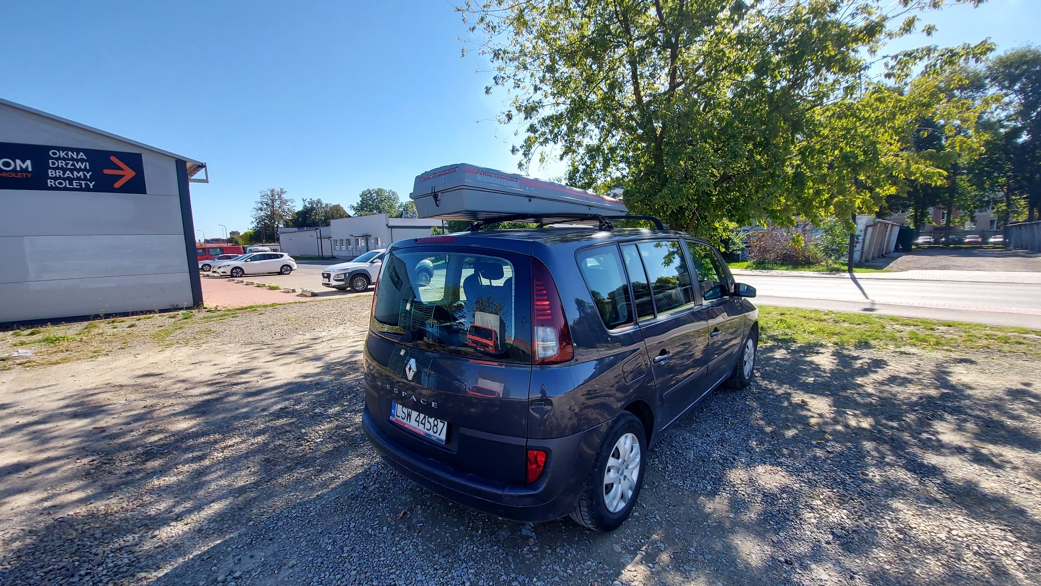 Granatowy Renault Espace z szarym boxem dachowym Thule na parkingu żwirowym, widok z tyłu, w tle budynek z reklamą okien i drzwi.