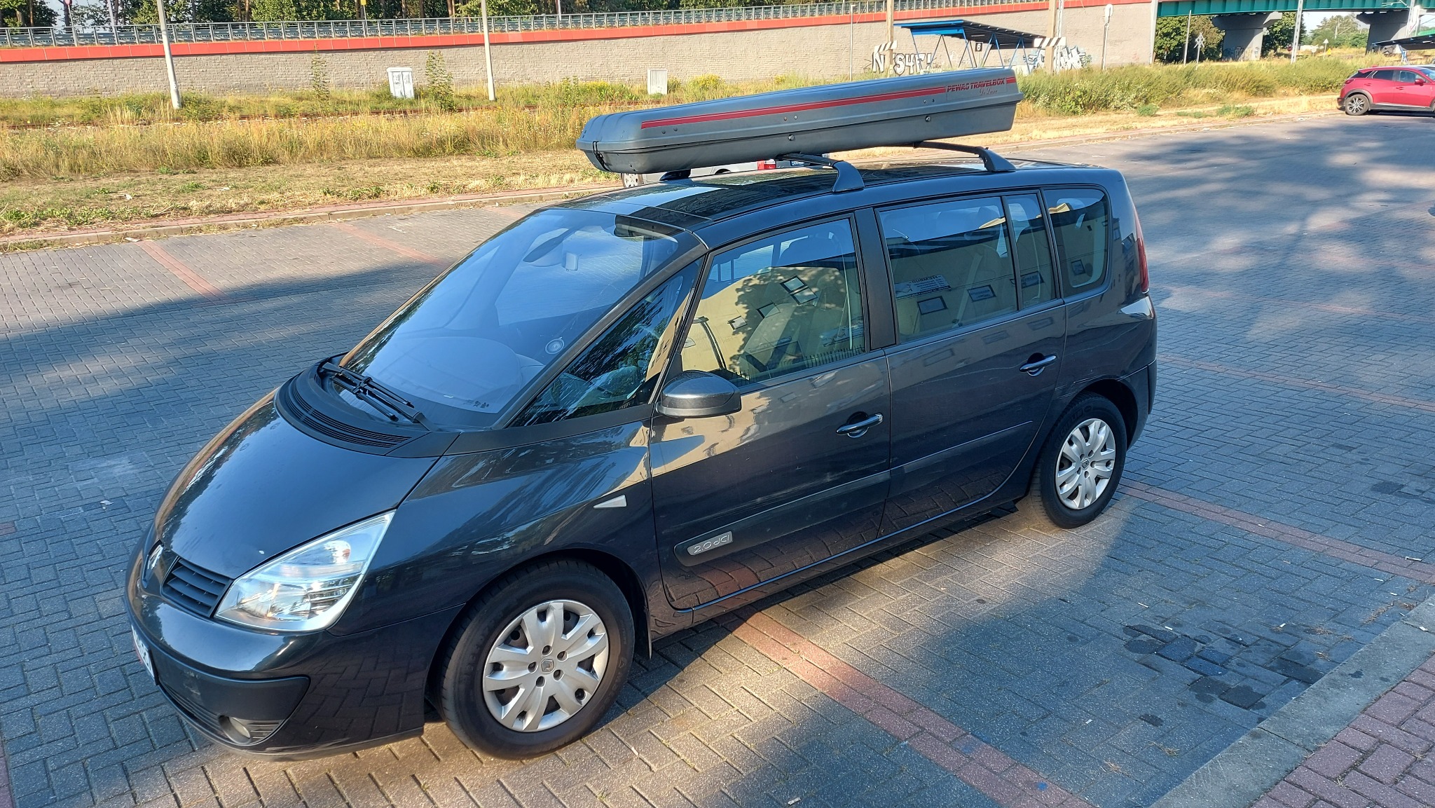 Ciemnoszary van Renault Espace z boxem dachowym na bagażniku, zaparkowany na brukowanej ulicy w słoneczny dzień.