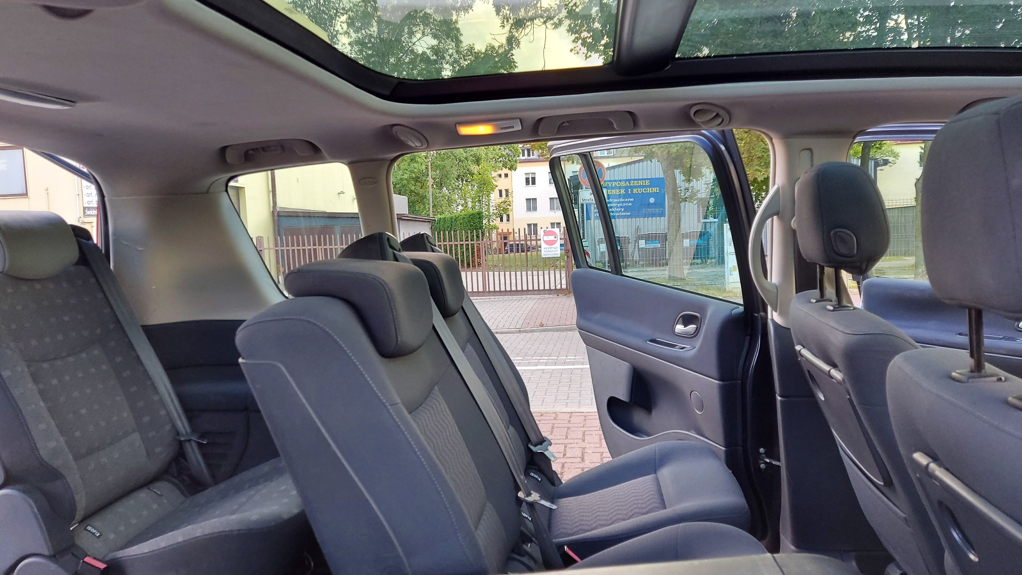 Wnętrze vana z otwartym dachem panoramicznym, widok na tylne siedzenia z zamontowanymi fotelikami ISOFIX i otwarte drzwi przesuwne, za którymi widać budynek z reklamą.