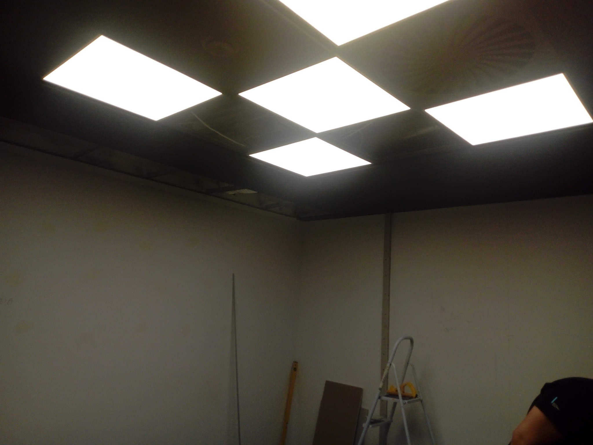 Sufit podwieszany z kwadratowymi panelami LED, drabina malarska i poziomnica oparta o ścianę w trakcie prac wykończeniowych.
