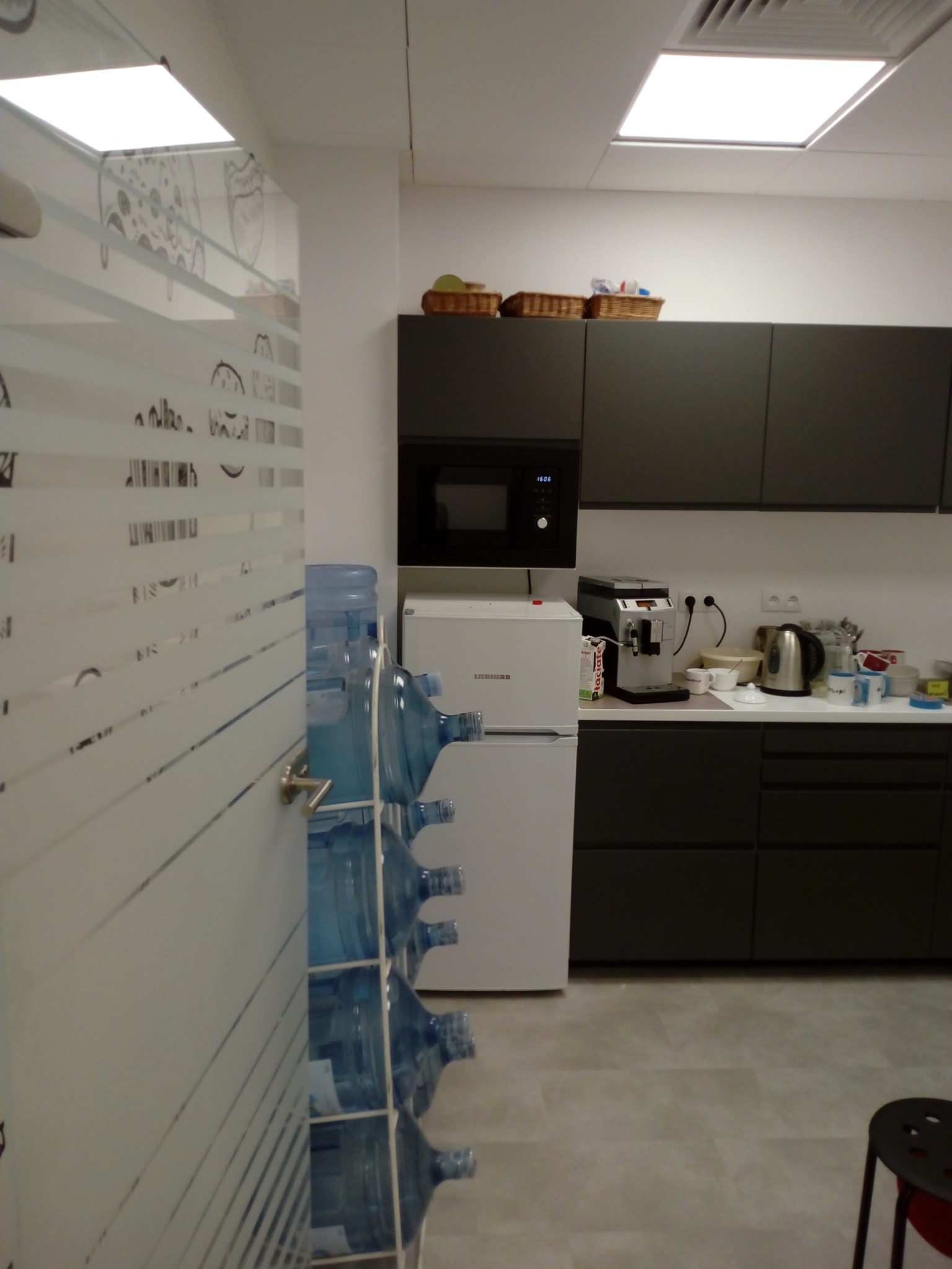 Wnętrze biurowej kuchni z ciemnymi szafkami, białymi sprzętami AGD i zapasem butli z wodą ustawionymi przy ścianie z dekoracyjną folią okienną.