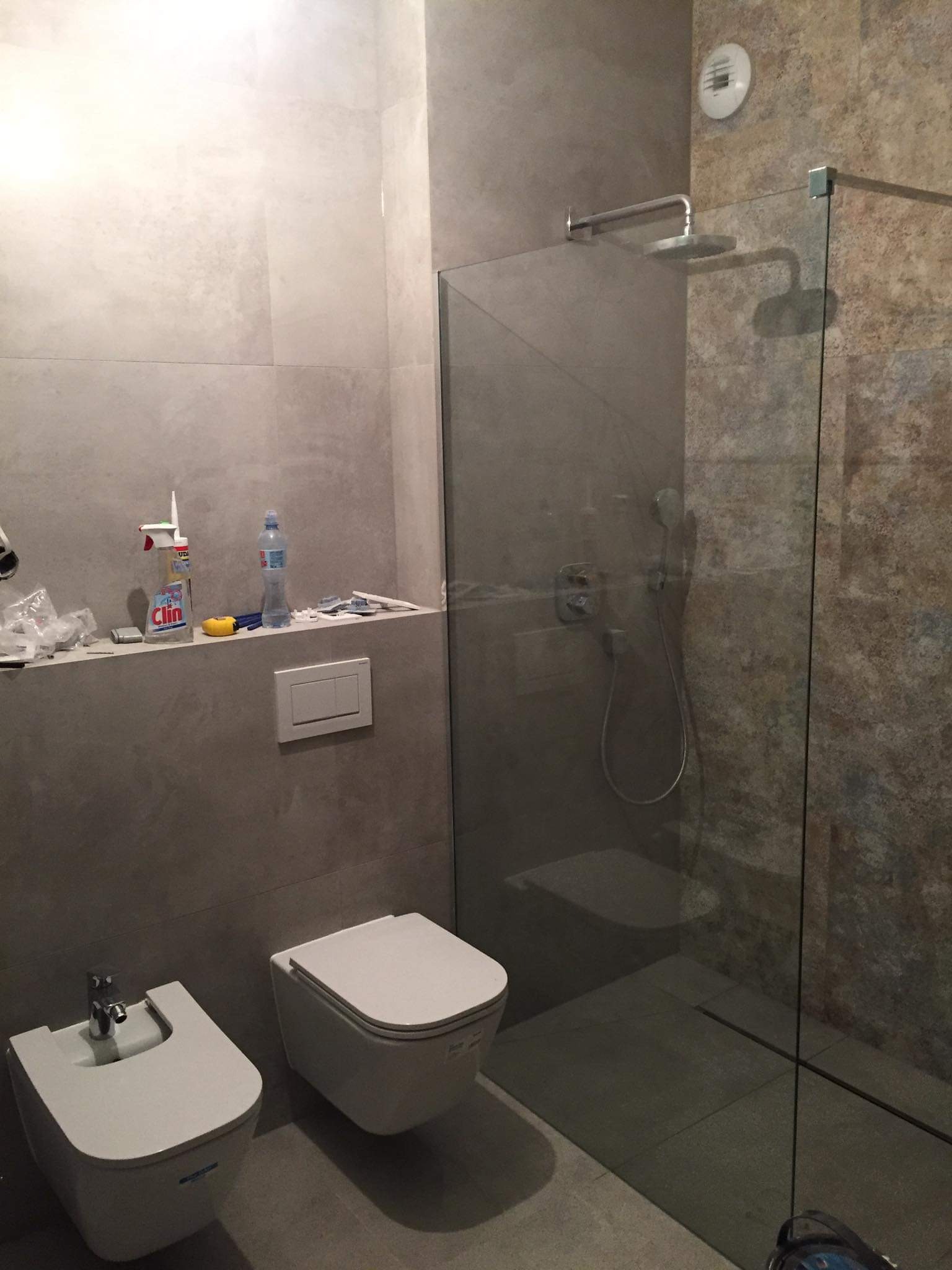 Nowoczesna łazienka z kabiną prysznicową typu walk-in, wiszącą toaletą i bidetem, wykończona płytkami w odcieniach szarości i beżu, z widocznymi akcesoriami i narzędziami na półce.