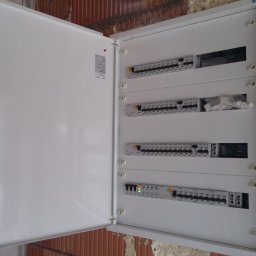 Proinstal - Otwarta biała skrzynka rozdzielcza z widocznymi bezpiecznikami i przewodami, zamontowana na surowej ceglanej ścianie w trakcie prac wykończeniowych.