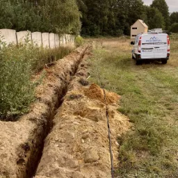 Wykopany rów w ziemi pod przyłącze elektryczne, widoczny kabel w czarnej osłonie, w tle zaparkowany biały samochód dostawczy z logo firmy Proinstal Sebastian Puchała Instalacje Elektryczne.