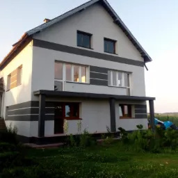 Dwupiętrowy dom z jasną elewacją i ciemnoszarymi pasami, widok z trawnika z zielenią, w tle pole kukurydzy.