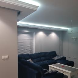 Ardbed Invest - Nowoczesny salon z narożnikiem w odcieniach granatu, oświetleniem LED w suficie i gładkimi, szarymi ścianami. Na suficie widoczne niedokończone podłączenie oświetlenia.