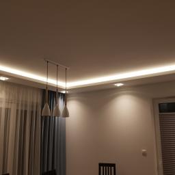 Ardbed Invest - Nowoczesne oświetlenie sufitowe z listwą LED i trzema wiszącymi lampami w jadalni, widok na okno z plisowaną roletą i zasłonami.