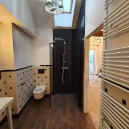 Nowoczesna łazienka z kabiną prysznicową wyłożoną czarnym marmurem, toaleta wisząca, grzejnik drabinkowy i drewnopodobna podłoga, oświetlona lampą wiszącą i światłem z okna dachowego.