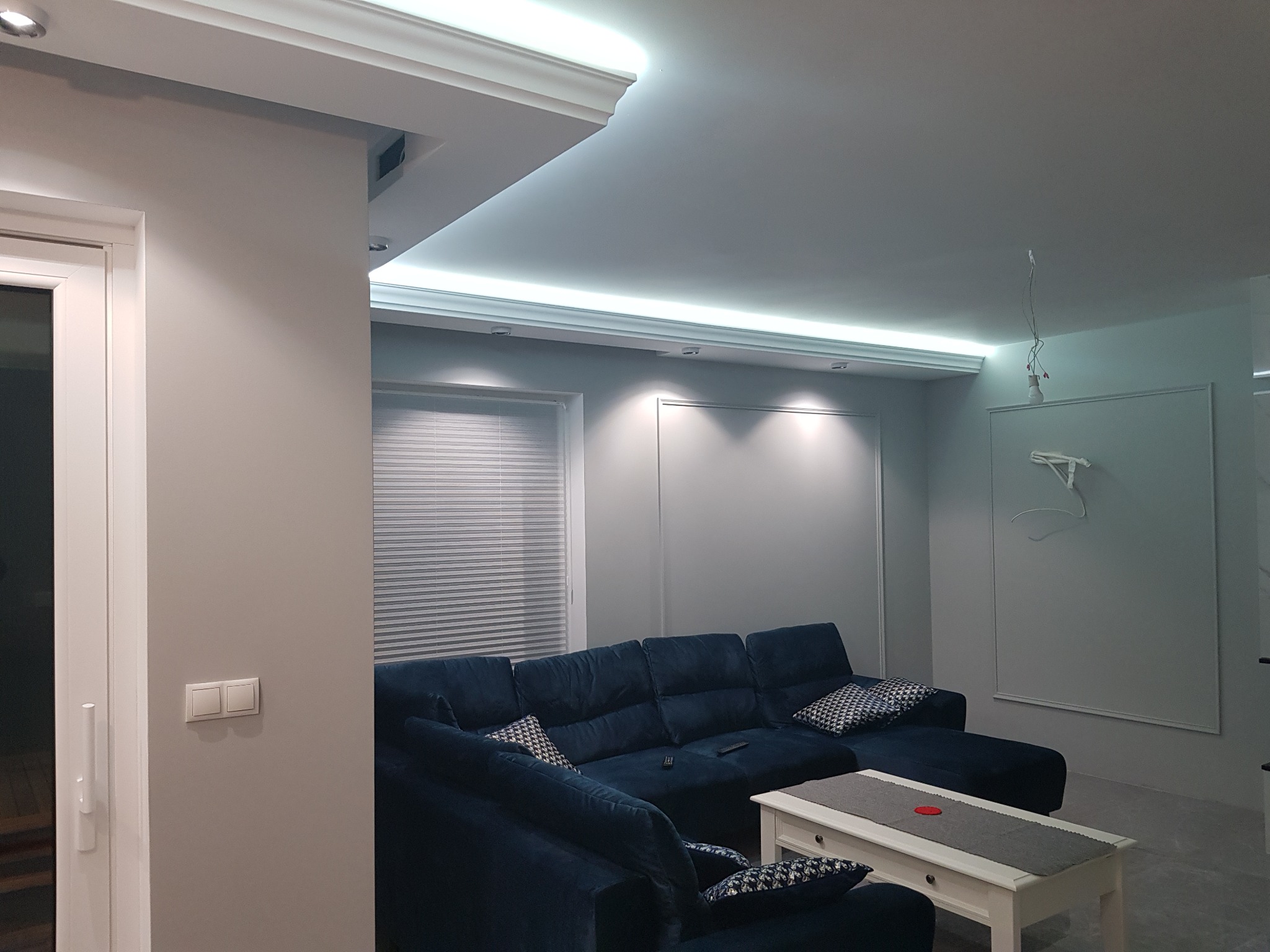 Nowoczesny salon z narożnikiem w odcieniach granatu, oświetleniem LED w suficie i gładkimi, szarymi ścianami. Na suficie widoczne niedokończone podłączenie oświetlenia.