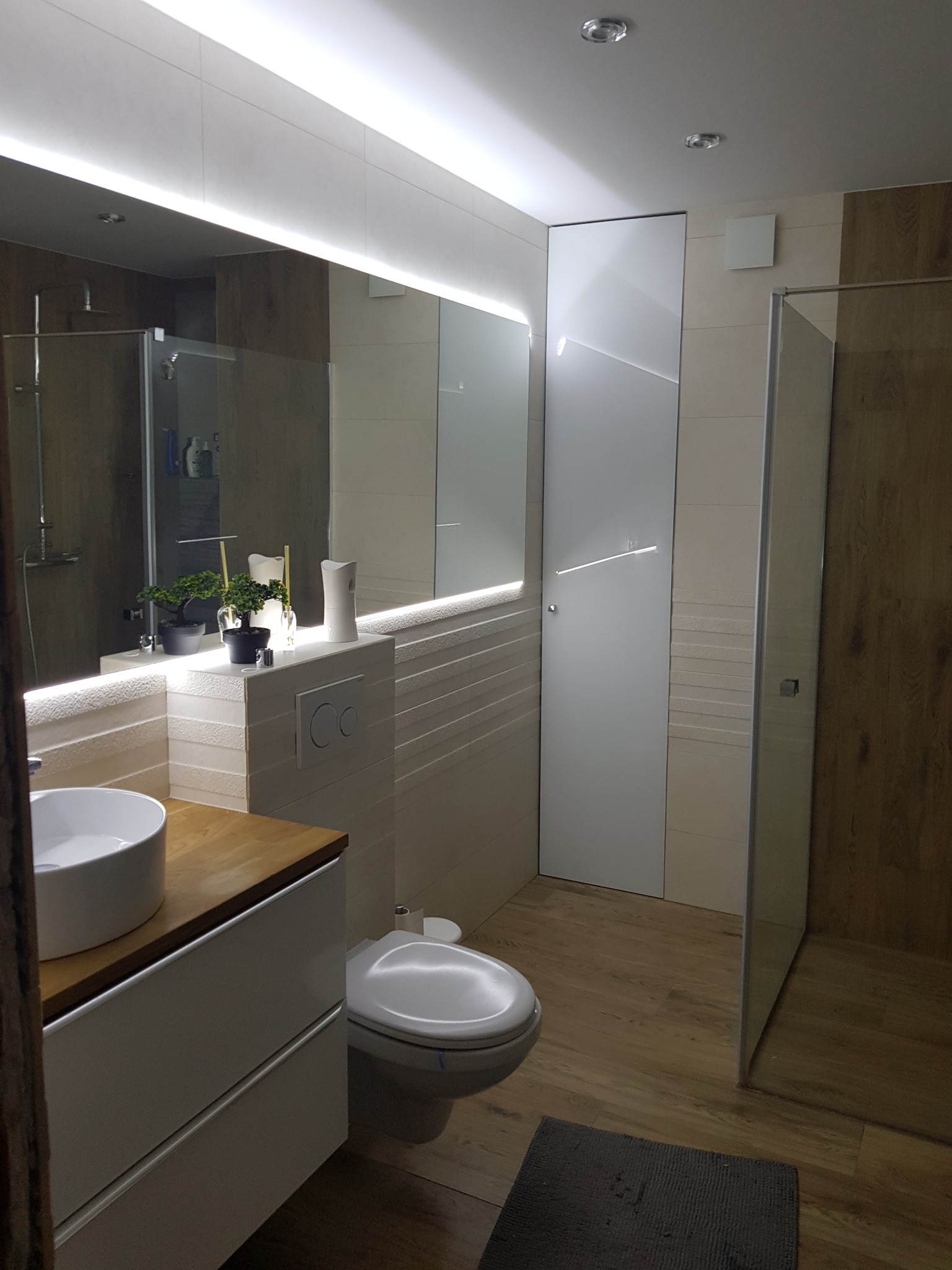 Nowoczesna łazienka z drewnianymi akcentami, podświetlanym lustrem i białymi płytkami o subtelnej fakturze, prezentująca minimalistyczny design z elementami roślinnymi na blacie.