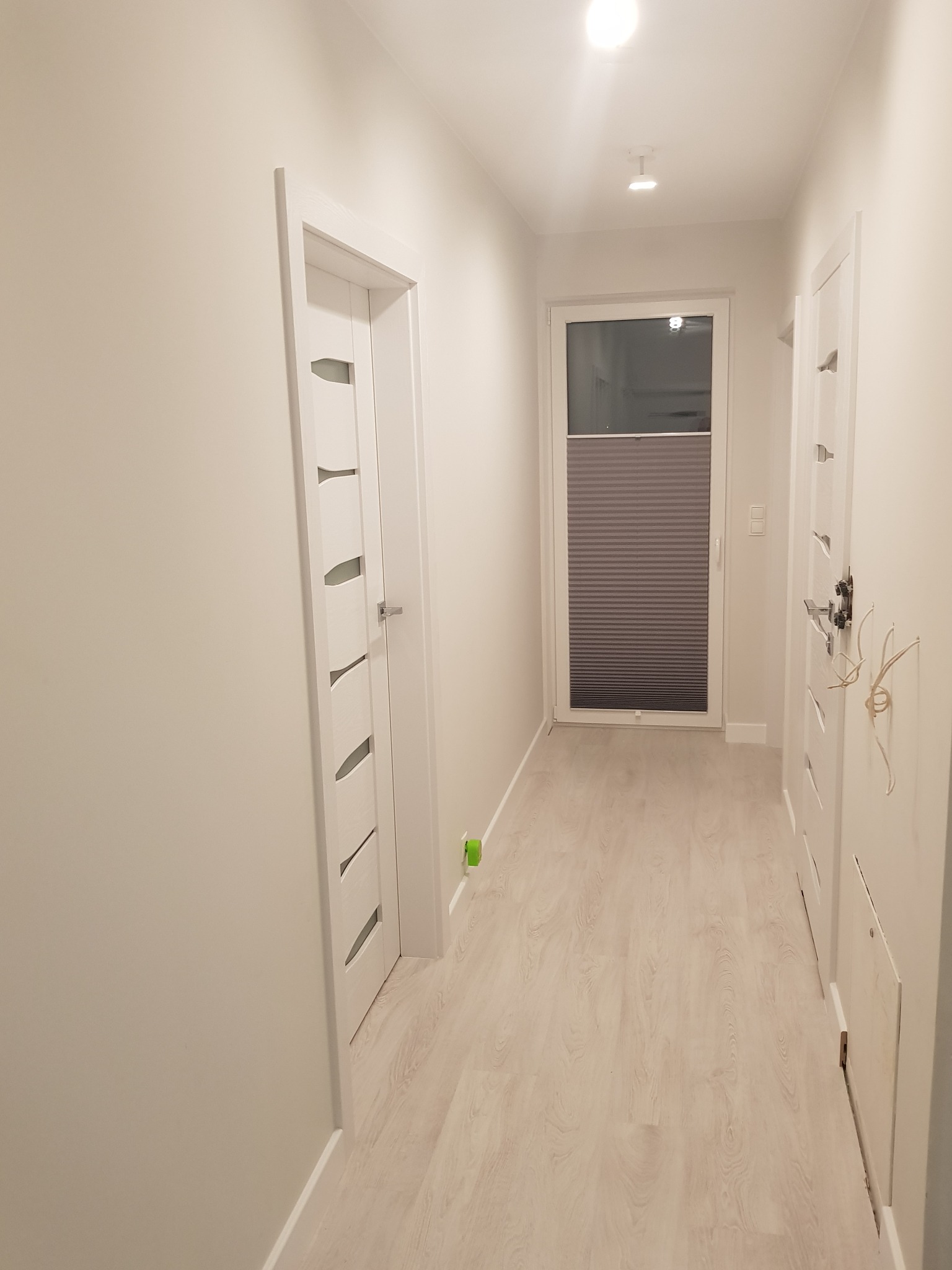 Jasny korytarz z białymi drzwiami z przeszkleniami, panelami podłogowymi i roletą plisowaną w oknie na końcu, widok z perspektywy.