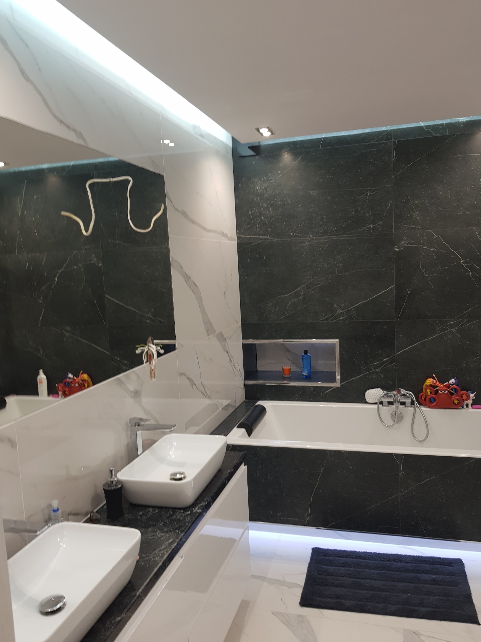 Nowoczesna łazienka z dwoma białymi umywalkami nablatowymi na blacie z czarnego marmuru, czarne marmurowe płytki na ścianach, podświetlana wnęka nad wanną, podświetlenie LED pod wanną i lustro...