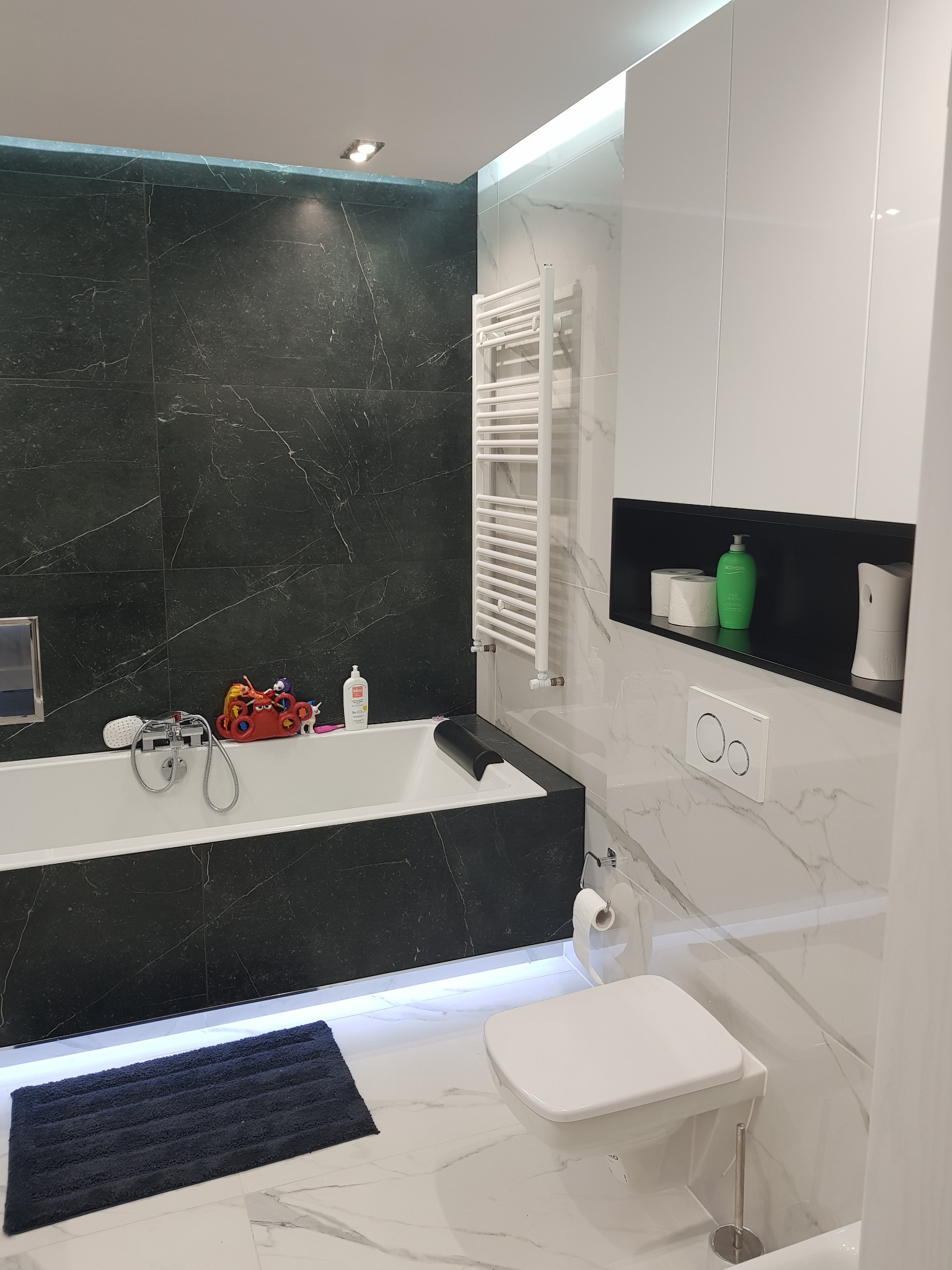 Nowoczesna łazienka z czarnymi i białymi marmurowymi płytkami, podświetlana wanna z zabawkami dla dzieci, grzejnik drabinkowy i toaleta wisząca.