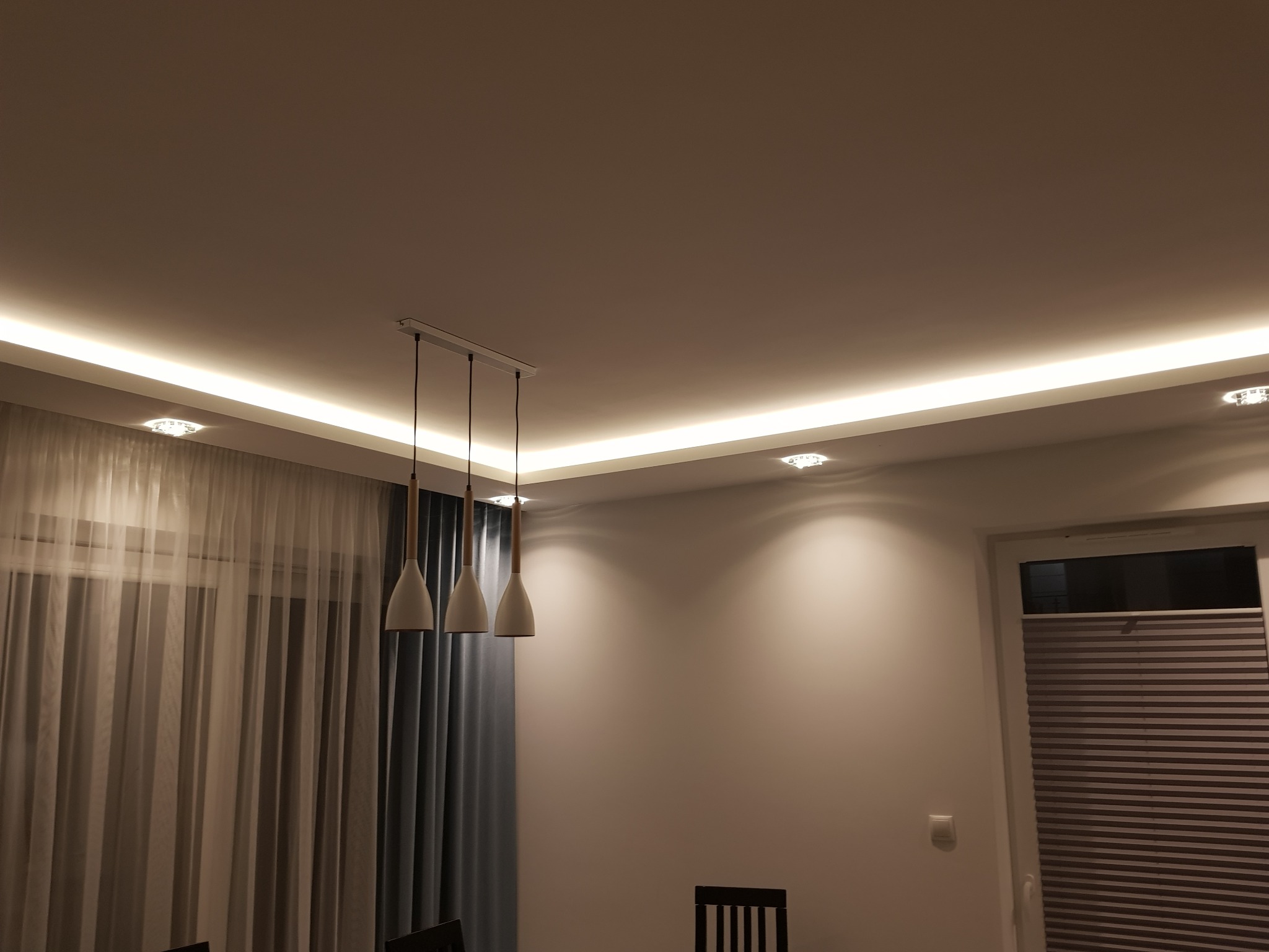 Nowoczesne oświetlenie sufitowe z listwą LED i trzema wiszącymi lampami w jadalni, widok na okno z plisowaną roletą i zasłonami.