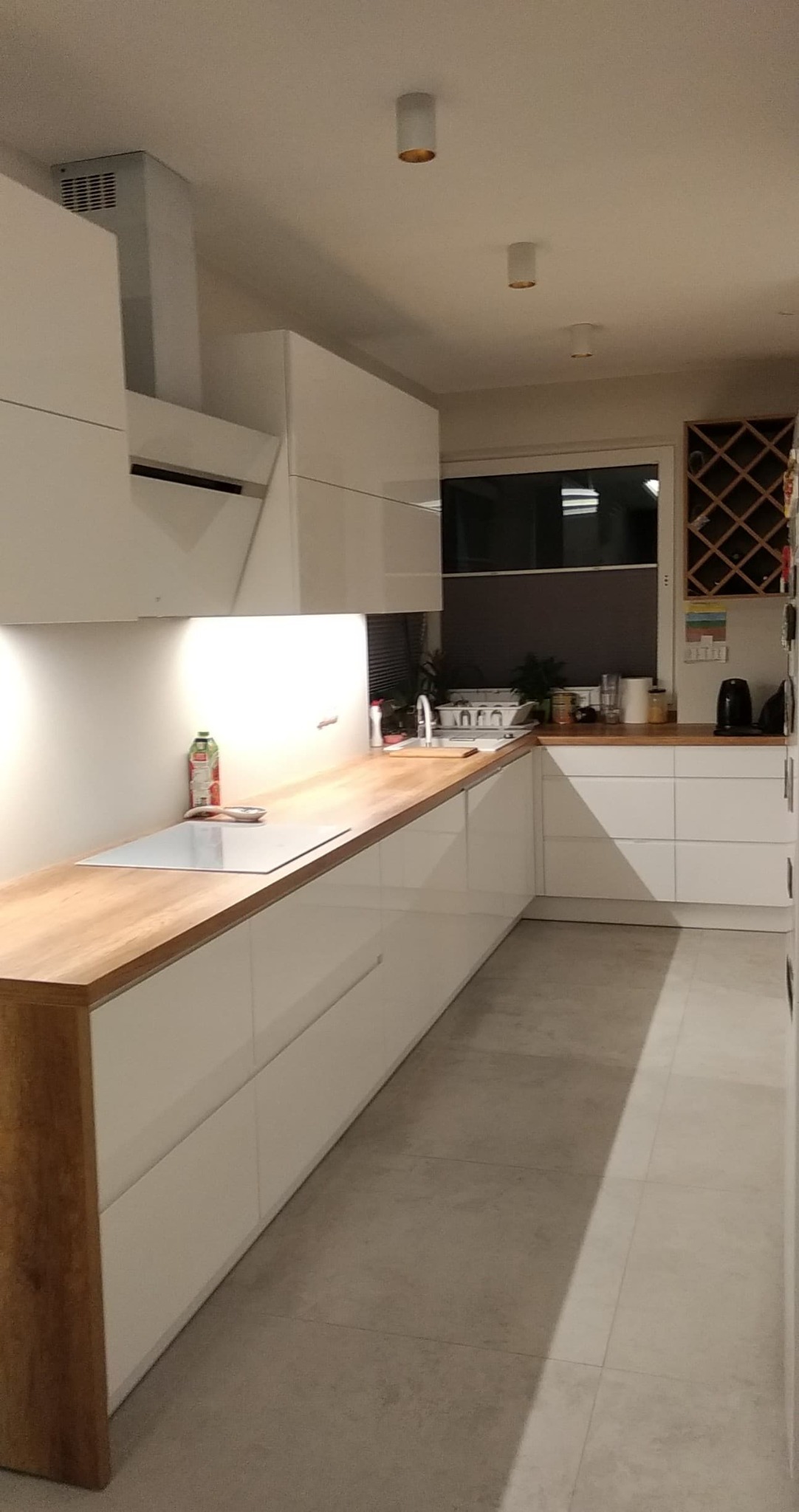 Nowoczesna kuchnia z białymi szafkami i drewnianym blatem, oświetlona punktowymi lampami sufitowymi, z widocznym okapem i półką na wino.