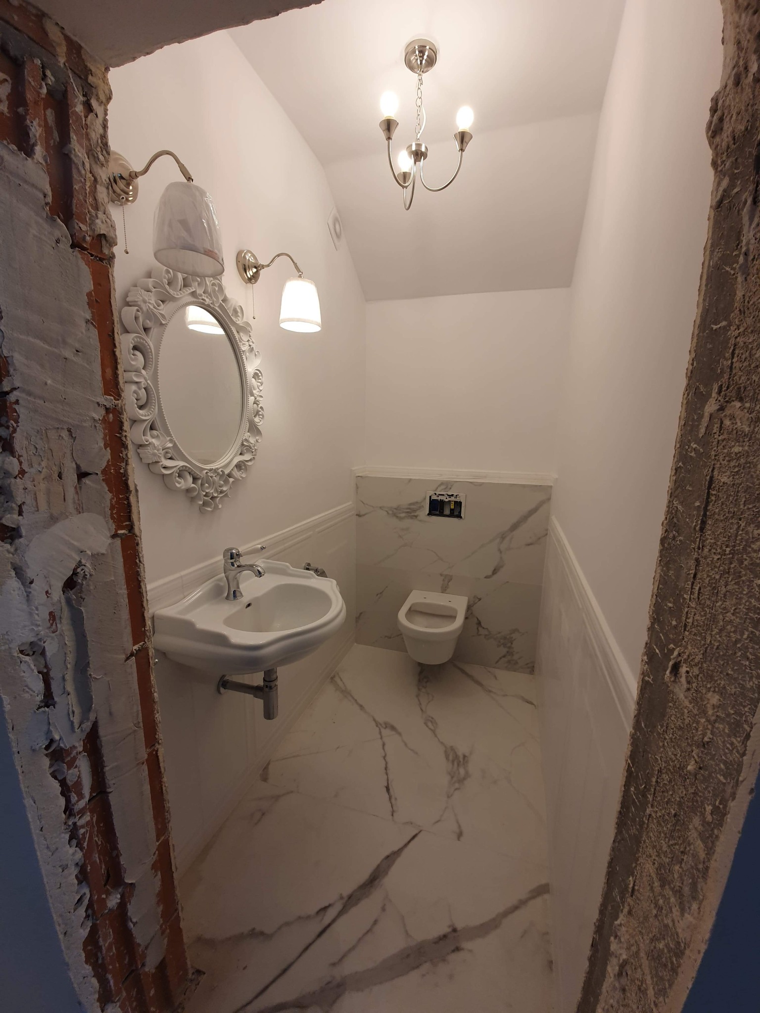 Biała łazienka z marmurowymi płytkami, widok na umywalkę z ozdobnym lustrem, toaletę i sufitową lampę w stylu retro, z widocznymi surowymi elementami ścian w trakcie remontu.
