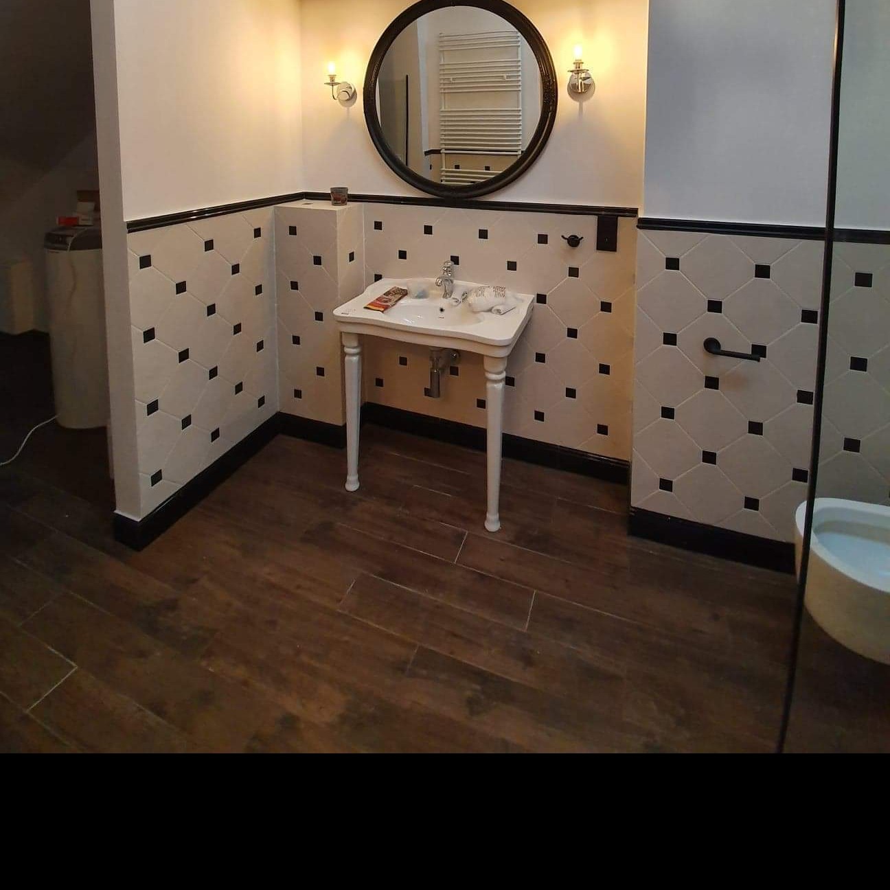 Ukończona łazienka z białą umywalką na nóżkach, okrągłym lustrem, drewnopodobną podłogą i ścianami wyłożonymi białymi płytkami z czarnymi kwadratami.