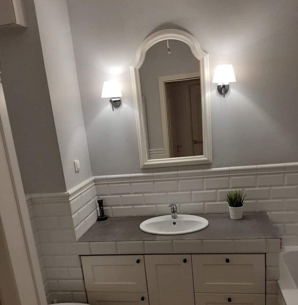 Jasna łazienka z owalną umywalką, białymi płytkami imitującymi cegłę, lustrem w ozdobnej ramie i kinkietami z abażurami.