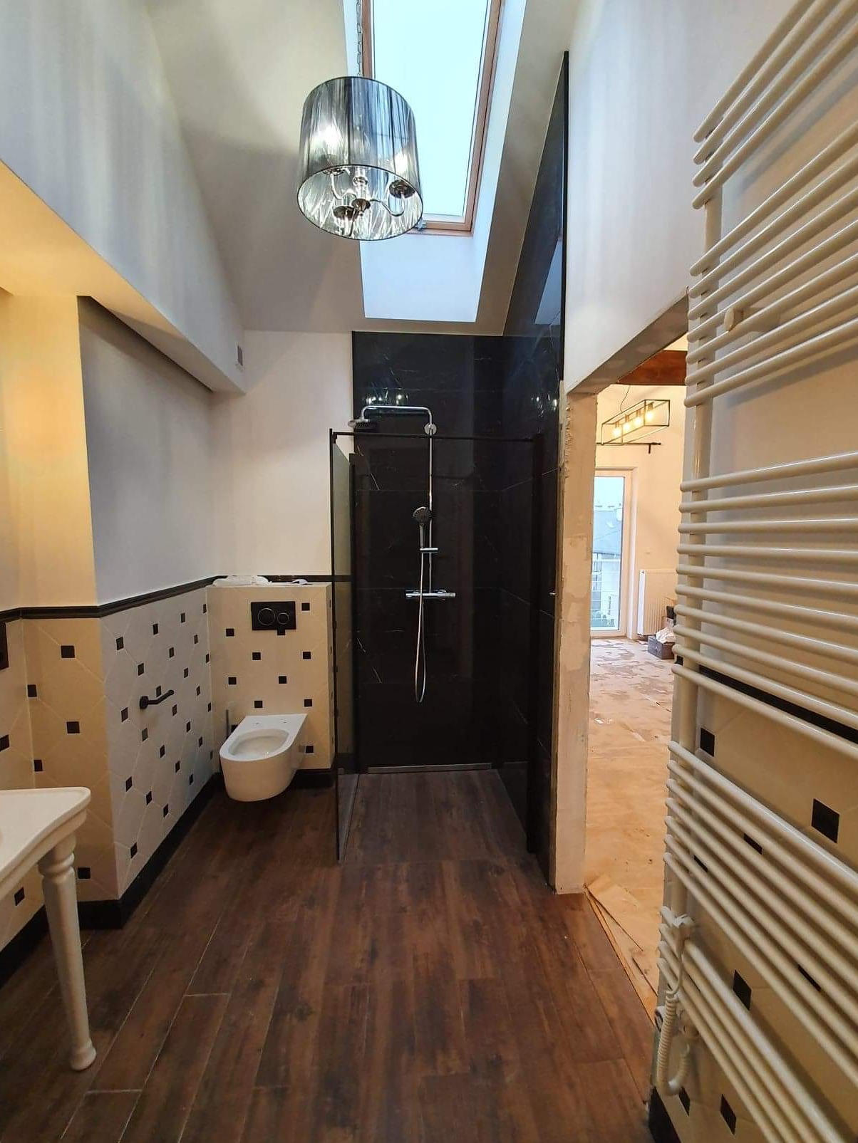 Nowoczesna łazienka z kabiną prysznicową wyłożoną czarnym marmurem, toaleta wisząca, grzejnik drabinkowy i drewnopodobna podłoga, oświetlona lampą wiszącą i światłem z okna dachowego.