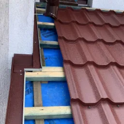 Częściowy widok dachu w trakcie montażu z widoczną konstrukcją drewnianą, membraną w kolorze niebieskim i częściowo ułożoną blachodachówką w kolorze bordowym oraz rynną spustową.