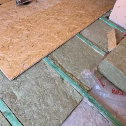 Płyta OSB częściowo ułożona na izolacji z wełny mineralnej pomiędzy legarami, w tle widoczne narzędzia: piła tarczowa i wkrętarka akumulatorowa.