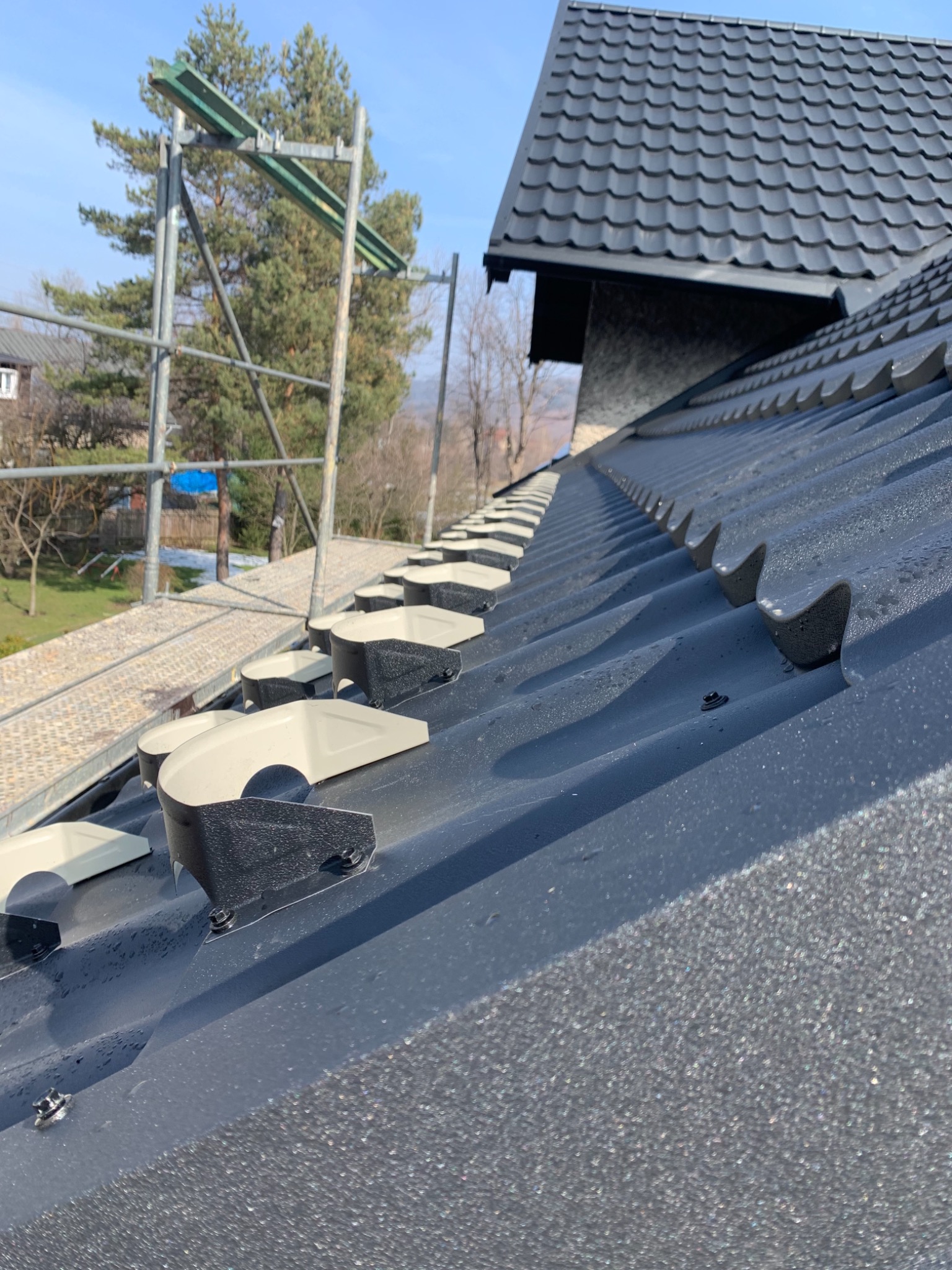 Nowy, grafitowy dach z zamontowanymi uchwytami rynnowymi i widocznymi kroplami wody, sfotografowany pod kątem ze stojącego rusztowania.