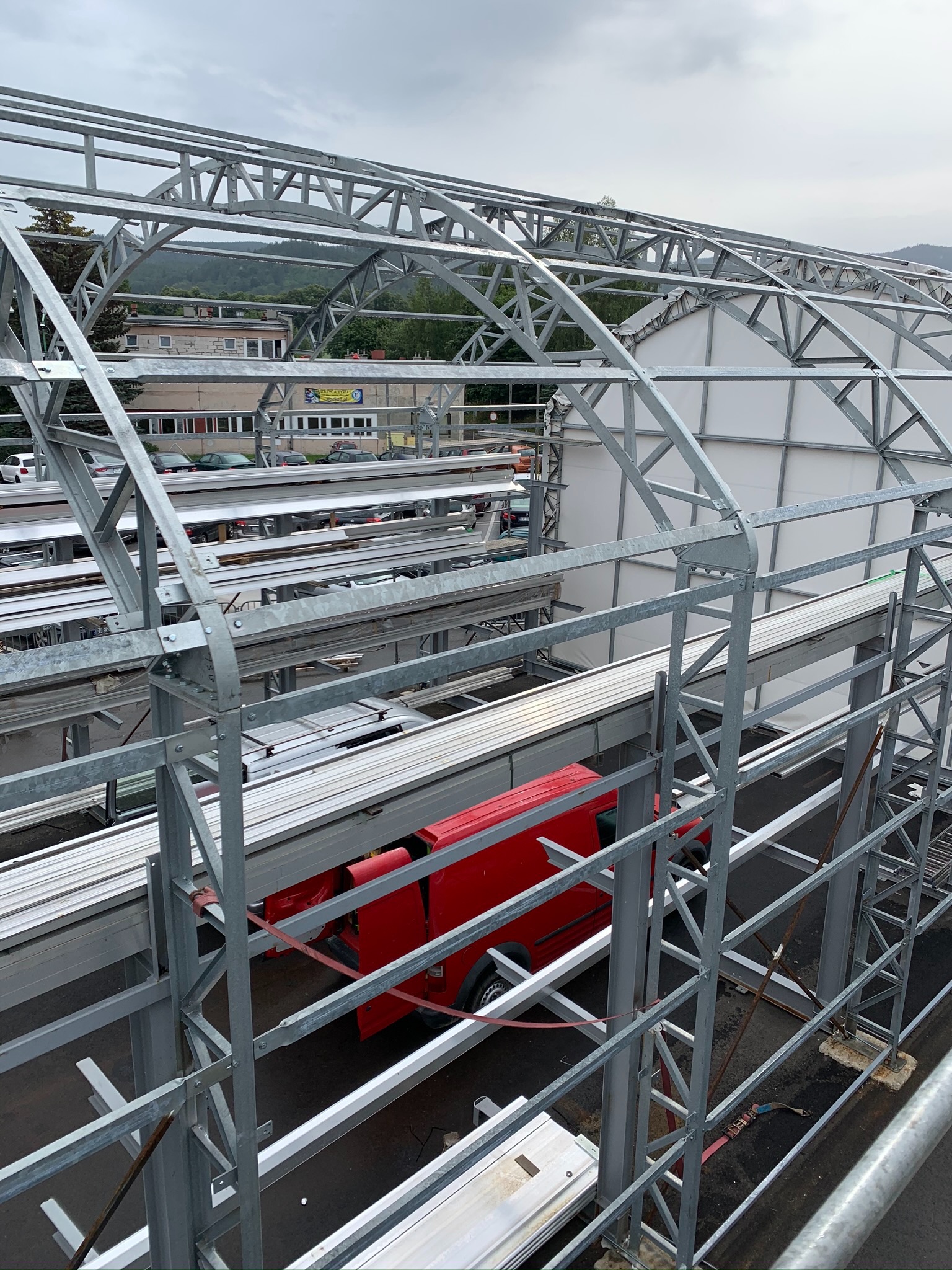 Metalowa konstrukcja szkieletowa w trakcie budowy, widoczny czerwony samochód dostawczy, w tle budynki i zalesione wzgórza pod pochmurnym niebem.