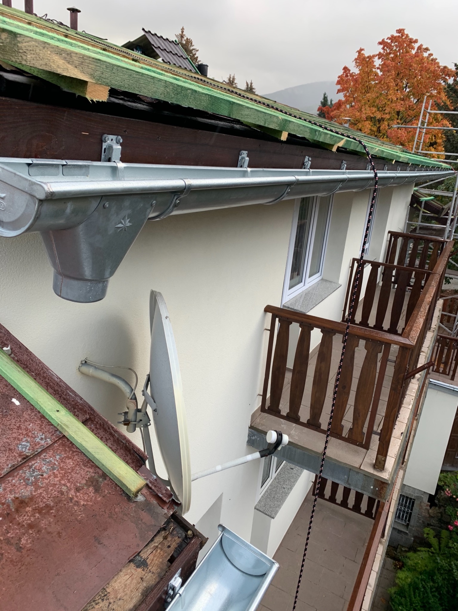 Szara rynna zamontowana na jasnej elewacji domu z drewnianym balkonem, widoczny fragment starego dachu i antena satelitarna, w tle drzewo z pomarańczowymi liśćmi i rusztowanie.