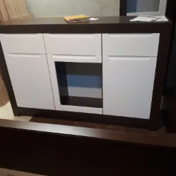 Biała komoda z ciemnobrązowymi elementami, widoczna w trakcie montażu, z drewnianymi elementami w tle.