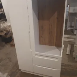 Biała szafa z dwoma szufladami i otwartą półką z drewnianym dekorem, widoczna w warsztacie podczas montażu.