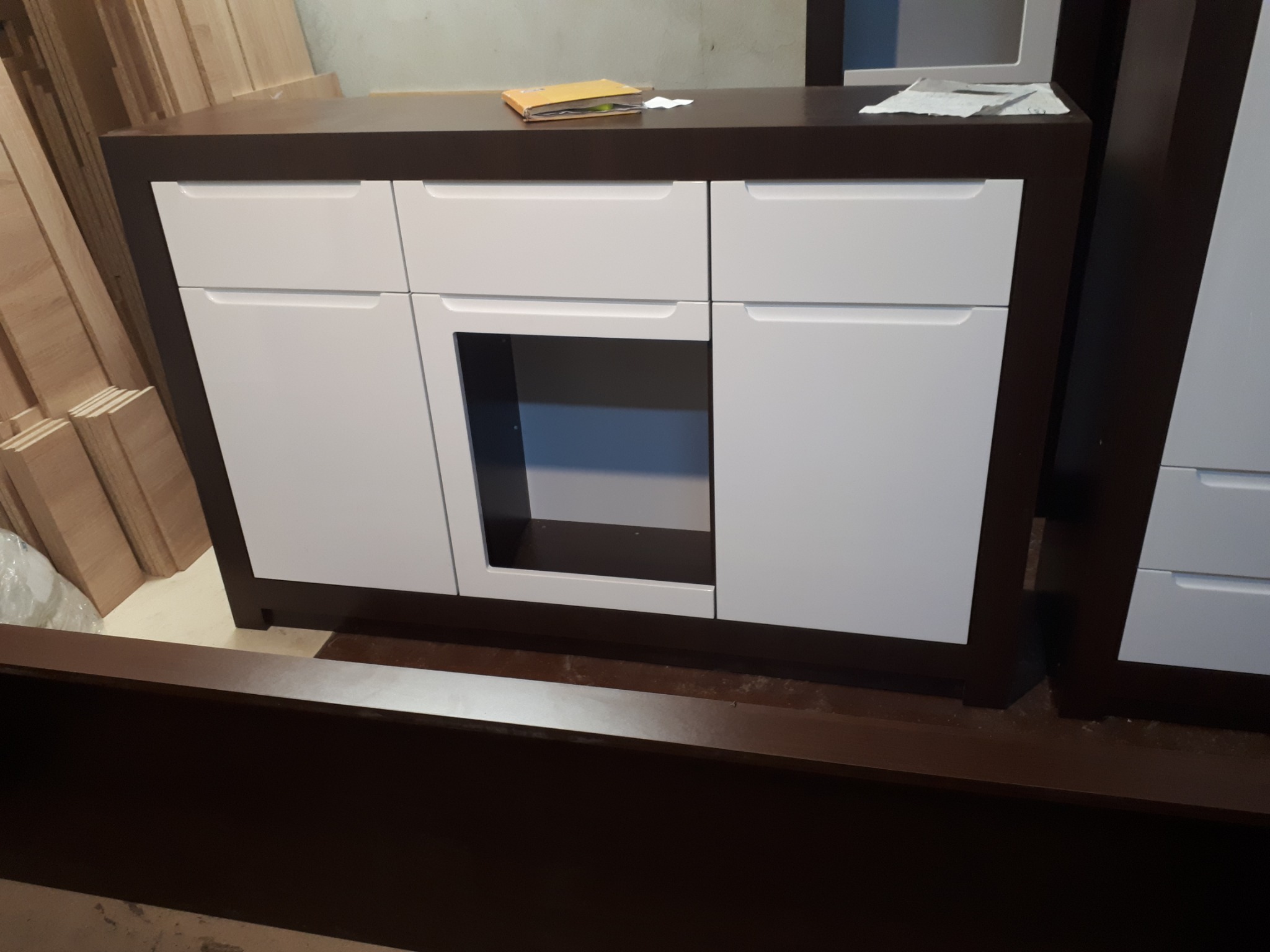 Biała komoda z ciemnobrązowymi elementami, widoczna w trakcie montażu, z drewnianymi elementami w tle.