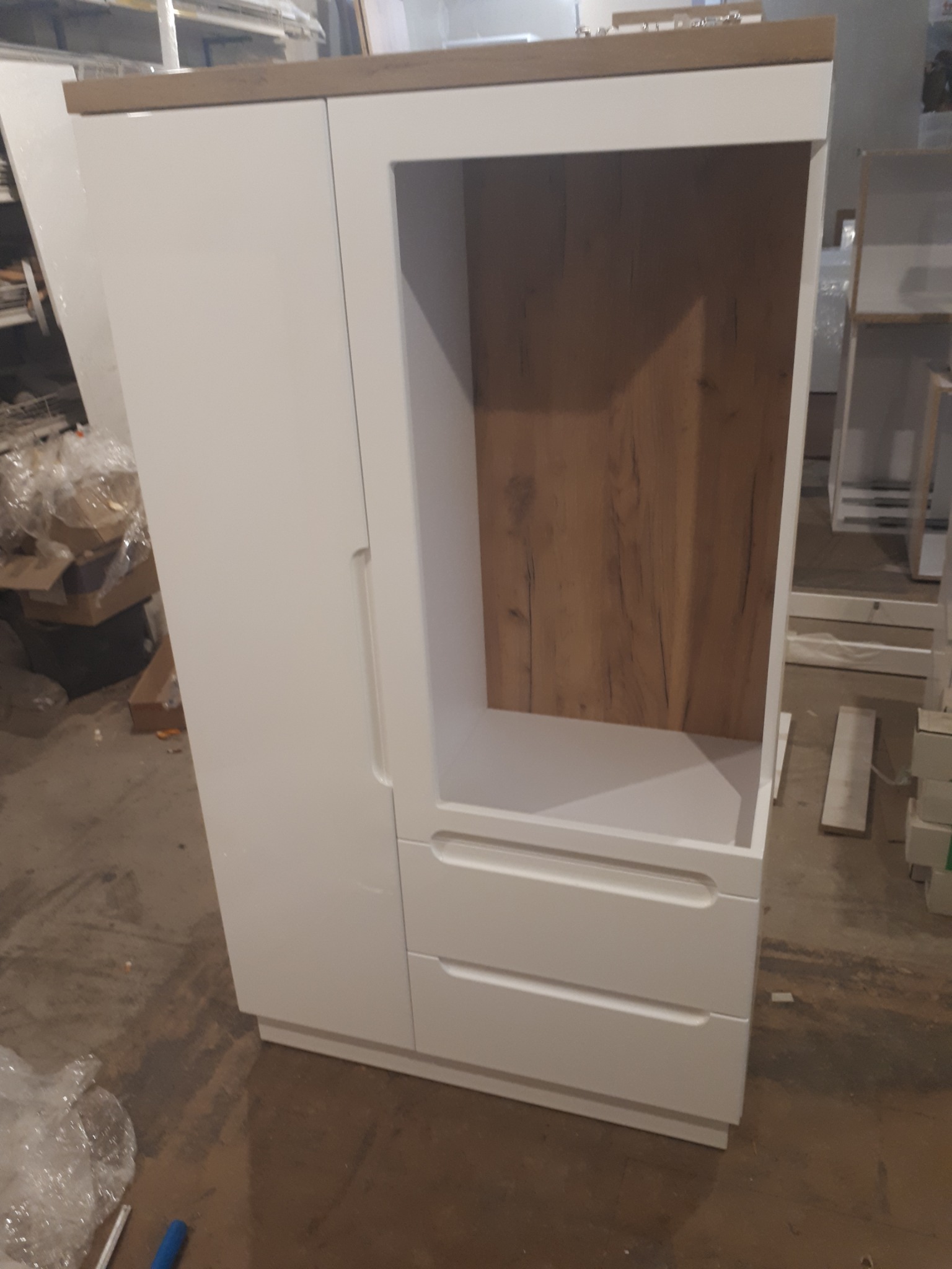 Biała szafa z dwoma szufladami i otwartą półką z drewnianym dekorem, widoczna w warsztacie podczas montażu.