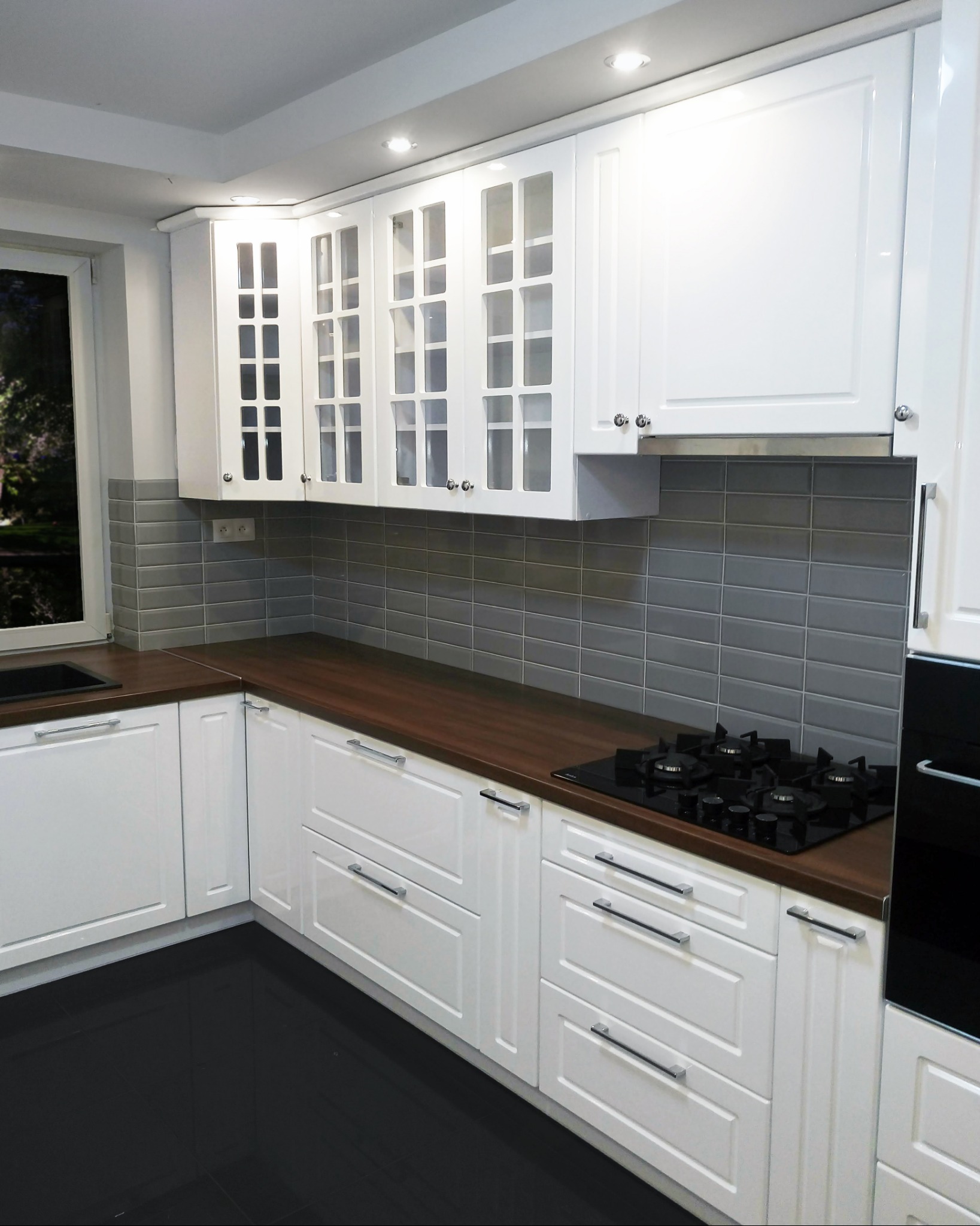 Nowoczesna, biała kuchnia z drewnianym blatem, szarymi płytkami i czarną płytą gazową, oświetlona punktowymi lampami.