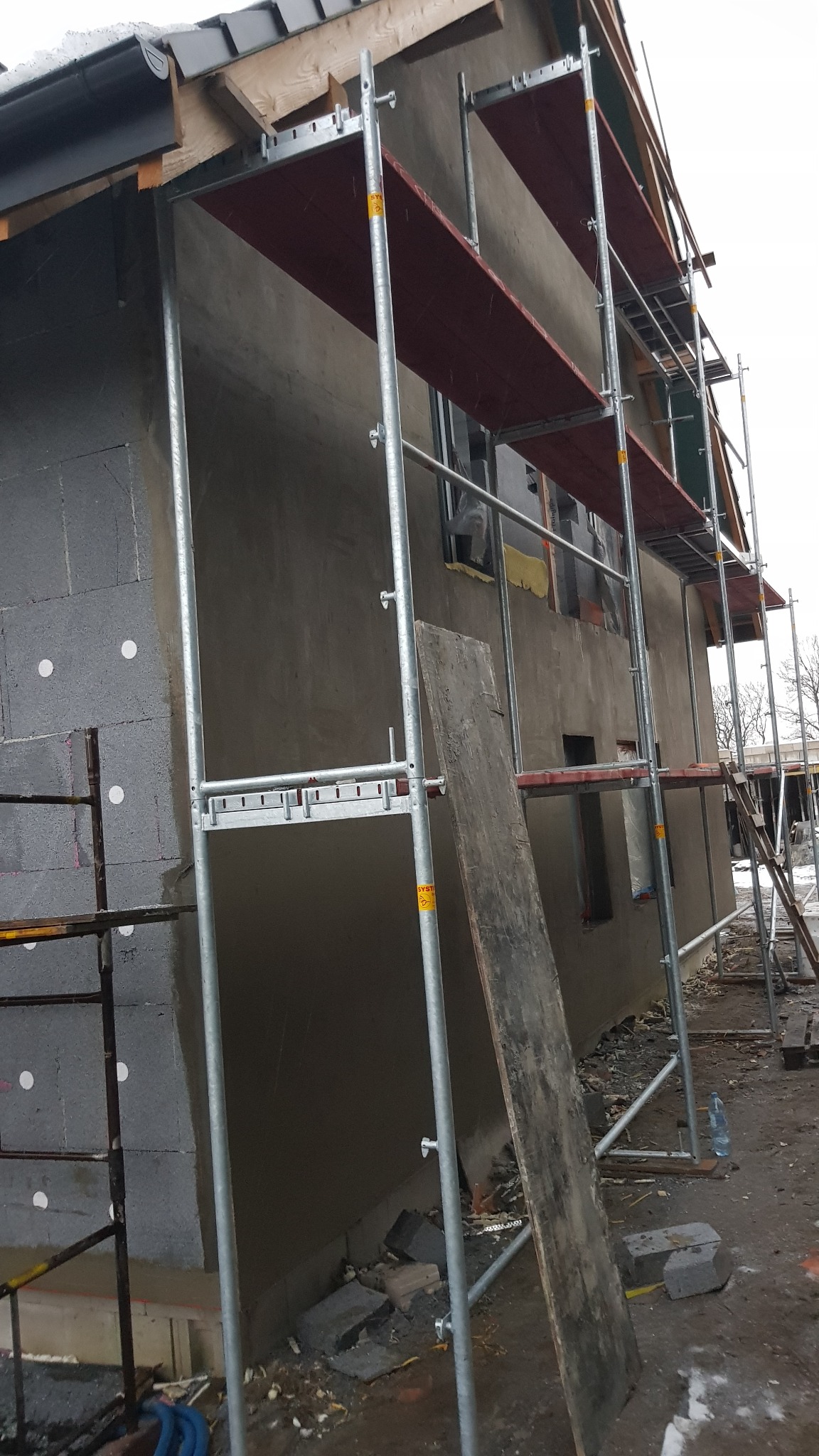 Budynek w trakcie ocieplania, widoczne rusztowania, styropian z kołkami oraz surowy tynk na elewacji bocznej, deska oparta o ścianę.