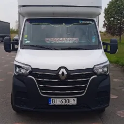 Przód białego busa Renault Master z wysoką kabiną, zaparkowanego na asfalcie z widocznymi śladami po malowaniu. Na przedniej szybie dekoracje i napis 'Polska'.