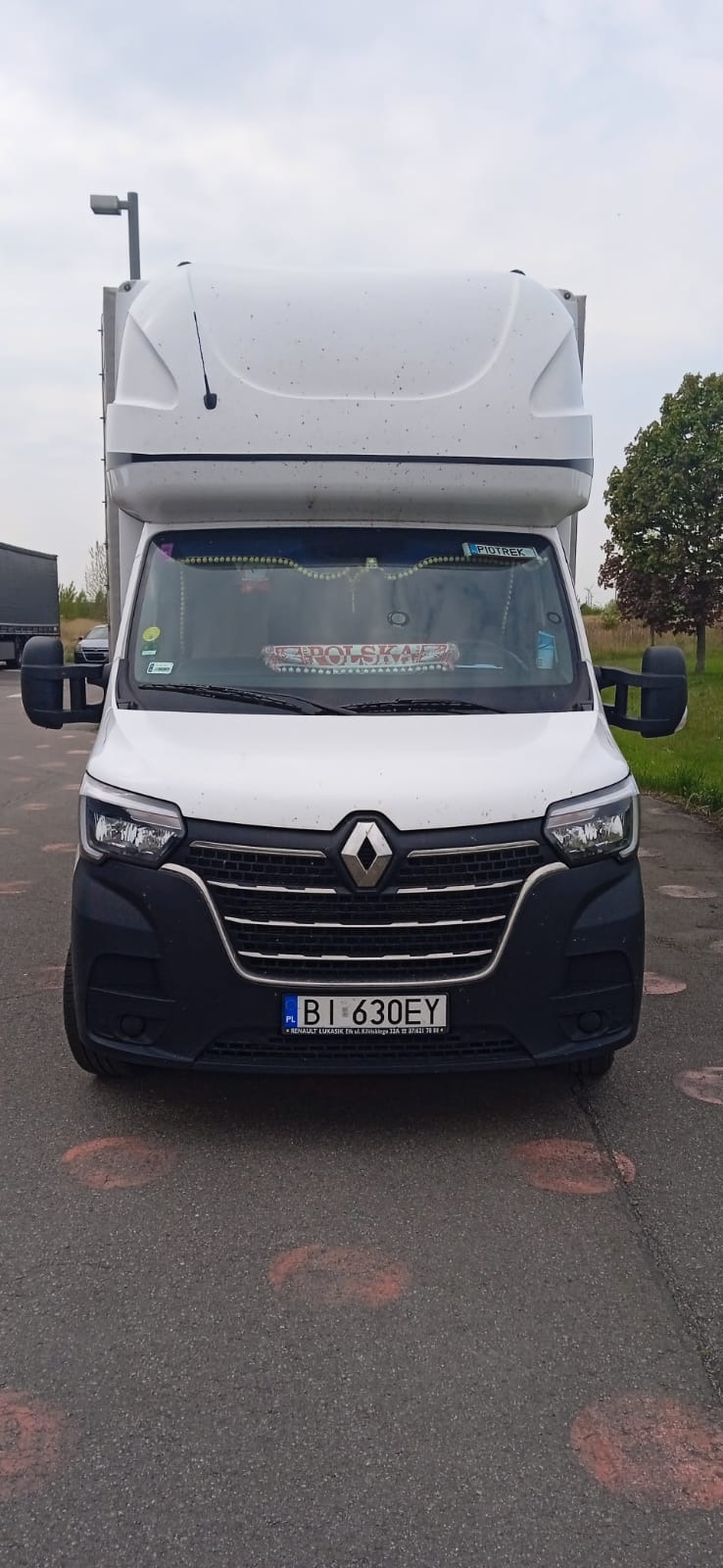 Przód białego busa Renault Master z wysoką kabiną, zaparkowanego na asfalcie z widocznymi śladami po malowaniu. Na przedniej szybie dekoracje i napis 'Polska'.