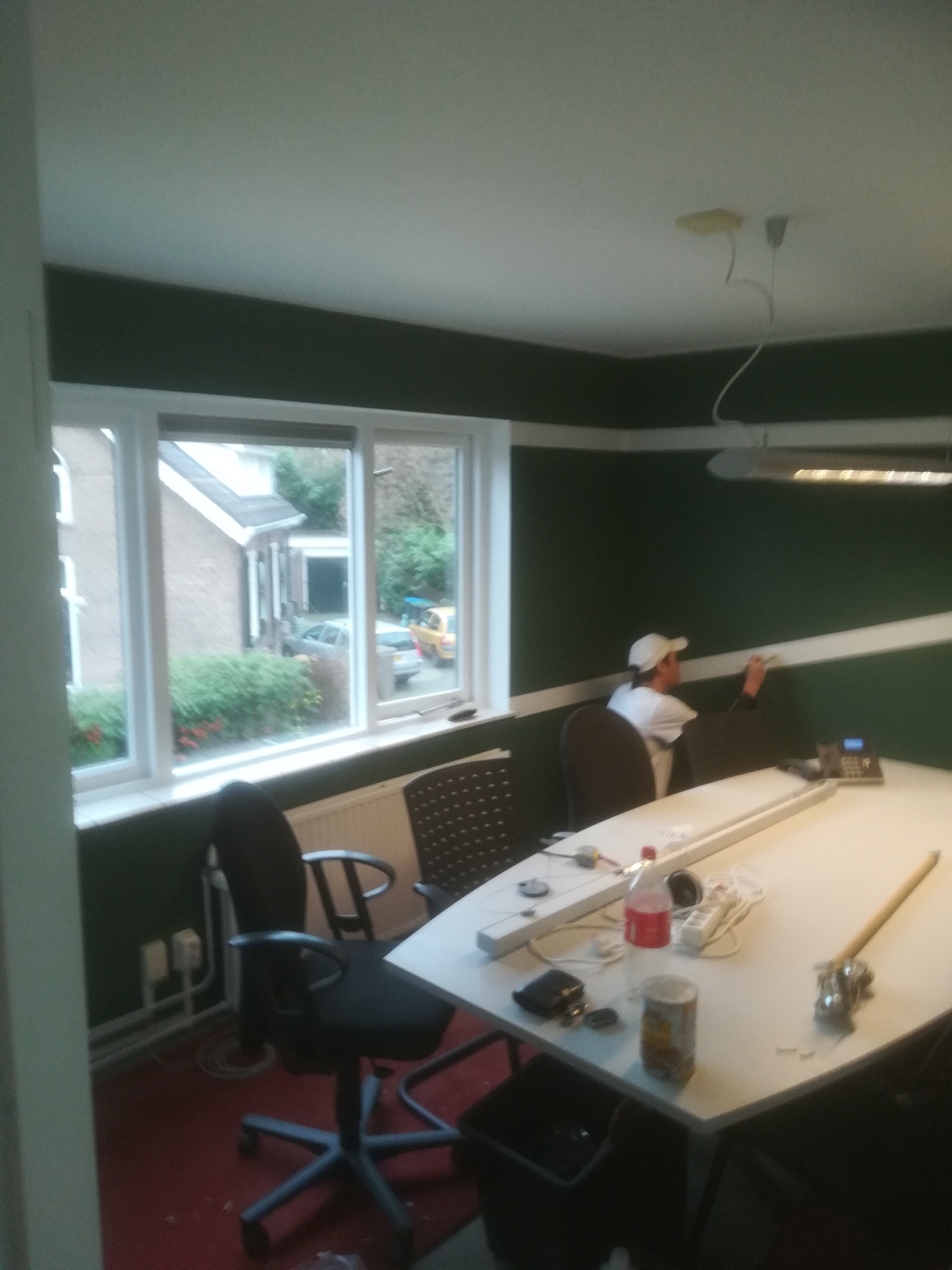 Mężczyzna w czapce maluje ciemnozieloną ścianę w pomieszczeniu z oknem, na stole widoczne narzędzia malarskie i napój.