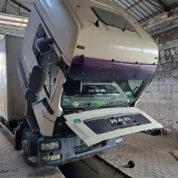 Ciężarówka MAN z podniesioną kabiną w warsztacie, widoczny silnik i kanał serwisowy w podłodze, obok ściana z cegieł i deski.