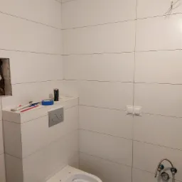 Biała łazienka w trakcie wykańczania, widoczny częściowo zamontowany sedes podtynkowy, białe płytki ścienne, wystające kable, otwór wentylacyjny oraz narzędzia i materiały budowlane na zabudowie...