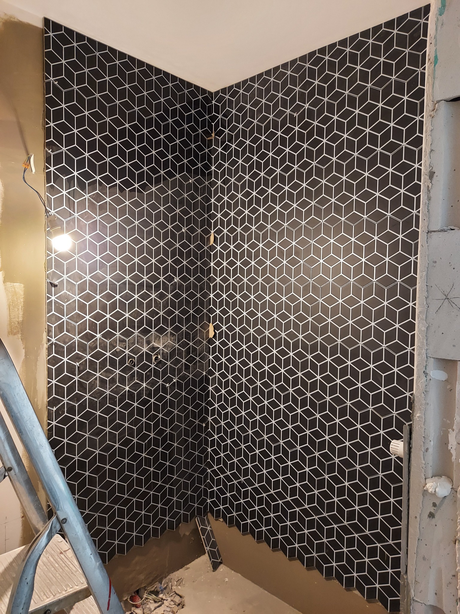 Fragment ściany łazienki w trakcie układania czarno-białych płytek z geometrycznym wzorem kostek 3D, widoczna drabina i niedokończone prace wykończeniowe.